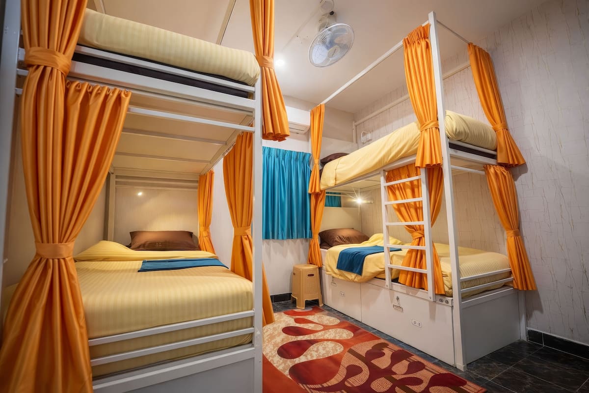 A dormitory room with bunk beds, orange curtains, a small stool, and a patterned rug.