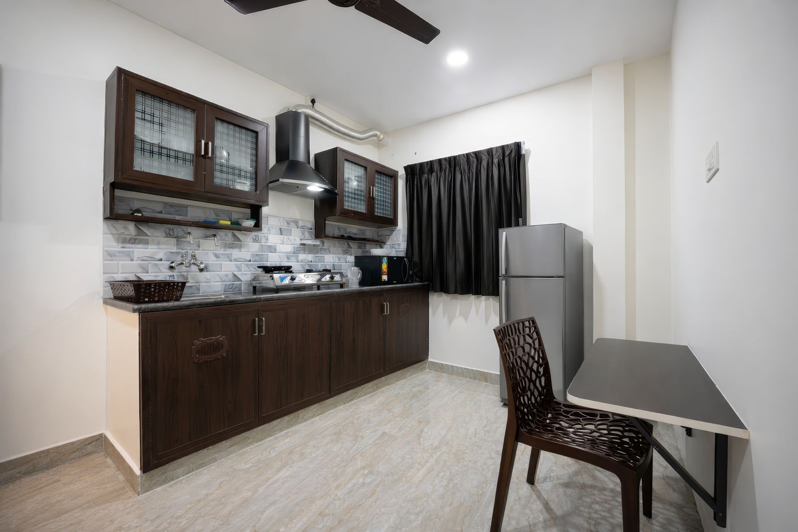 A compact kitchen with dark wooden cabinets, a stainless steel refrigerator, and a dining area with a simple table and chair.