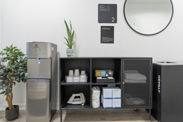 A kitchenette with a water dispenser, a potted plant, a shelf with cups and containers, a trash bin, and a wall-mounted sign indicating room number 2.