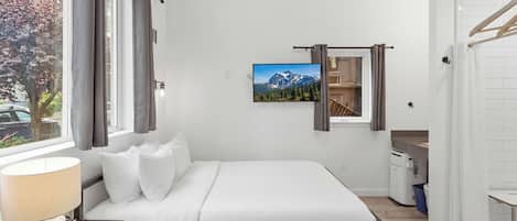 A bedroom with a bed, a TV, a window with curtains, a lamp, and a view of mountains.