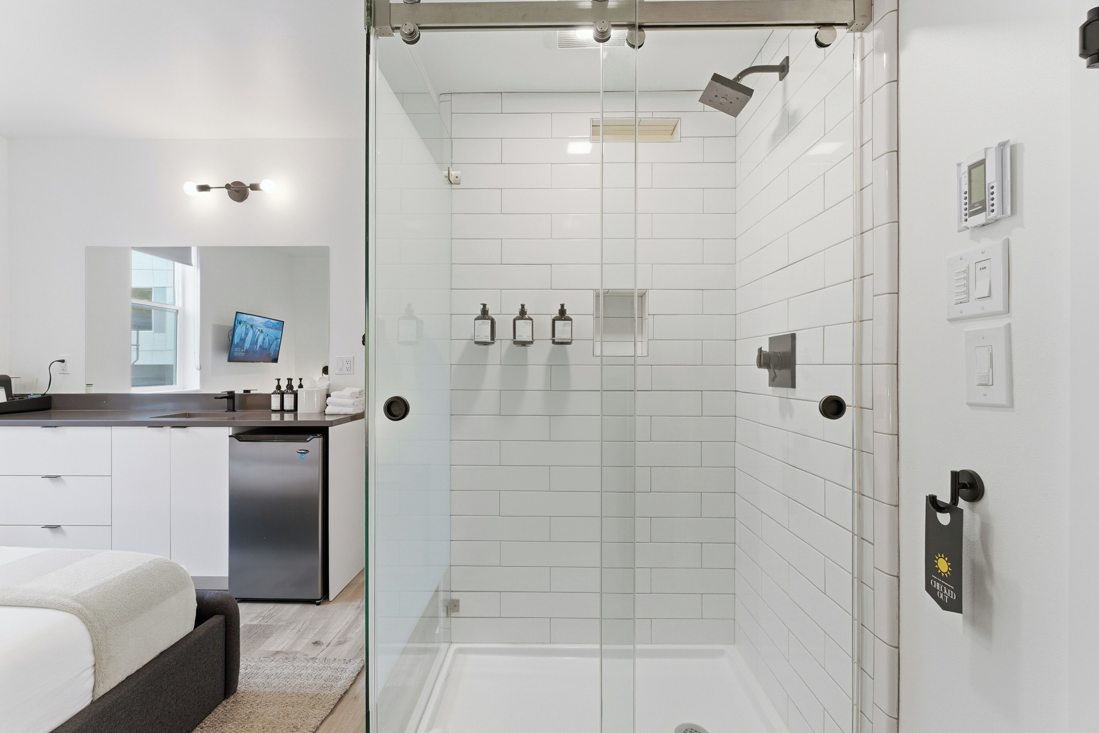 Un baño moderno con cabina de ducha de vidrio, paredes con azulejos blancos, lavamanos empotrado y un televisor de pantalla plana sobre el espejo.