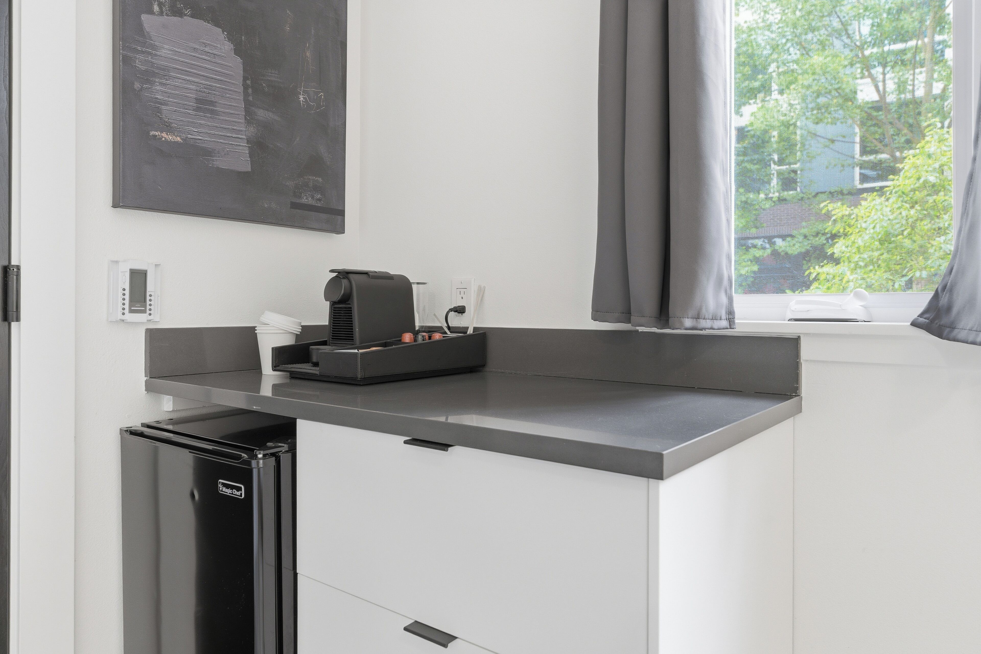 A kitchenette with a coffee machine, a small refrigerator, and a window with curtains.