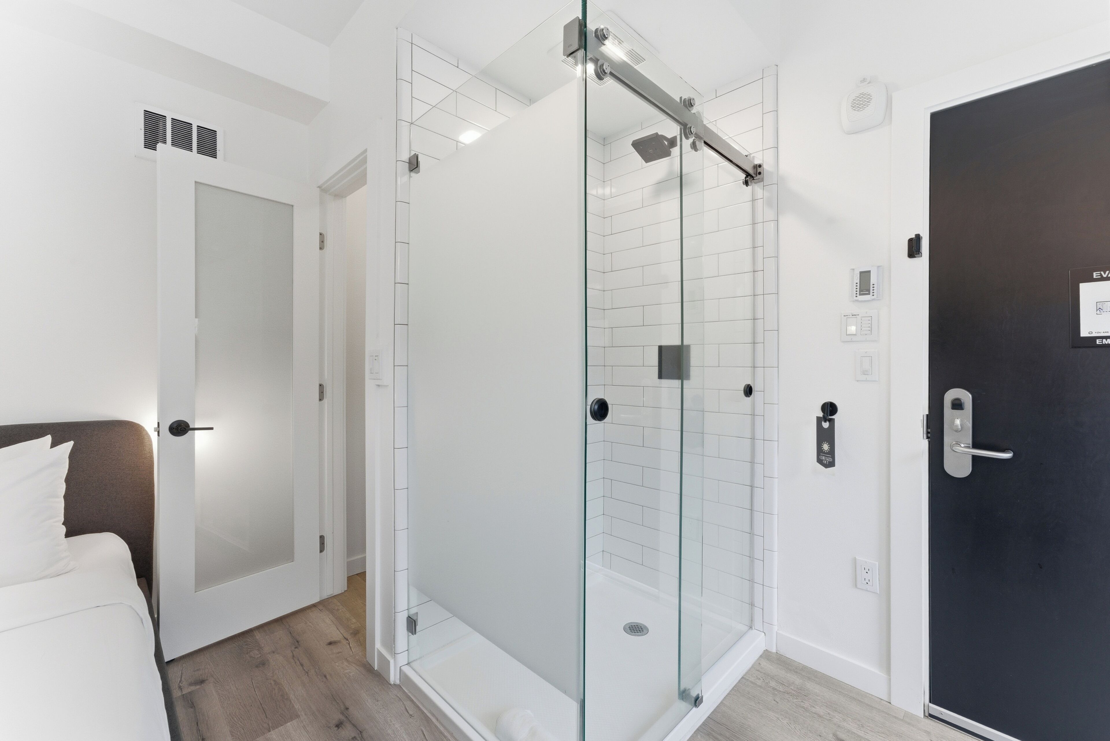A modern bathroom with a glass-enclosed shower, a white door, and a bed with white bedding.