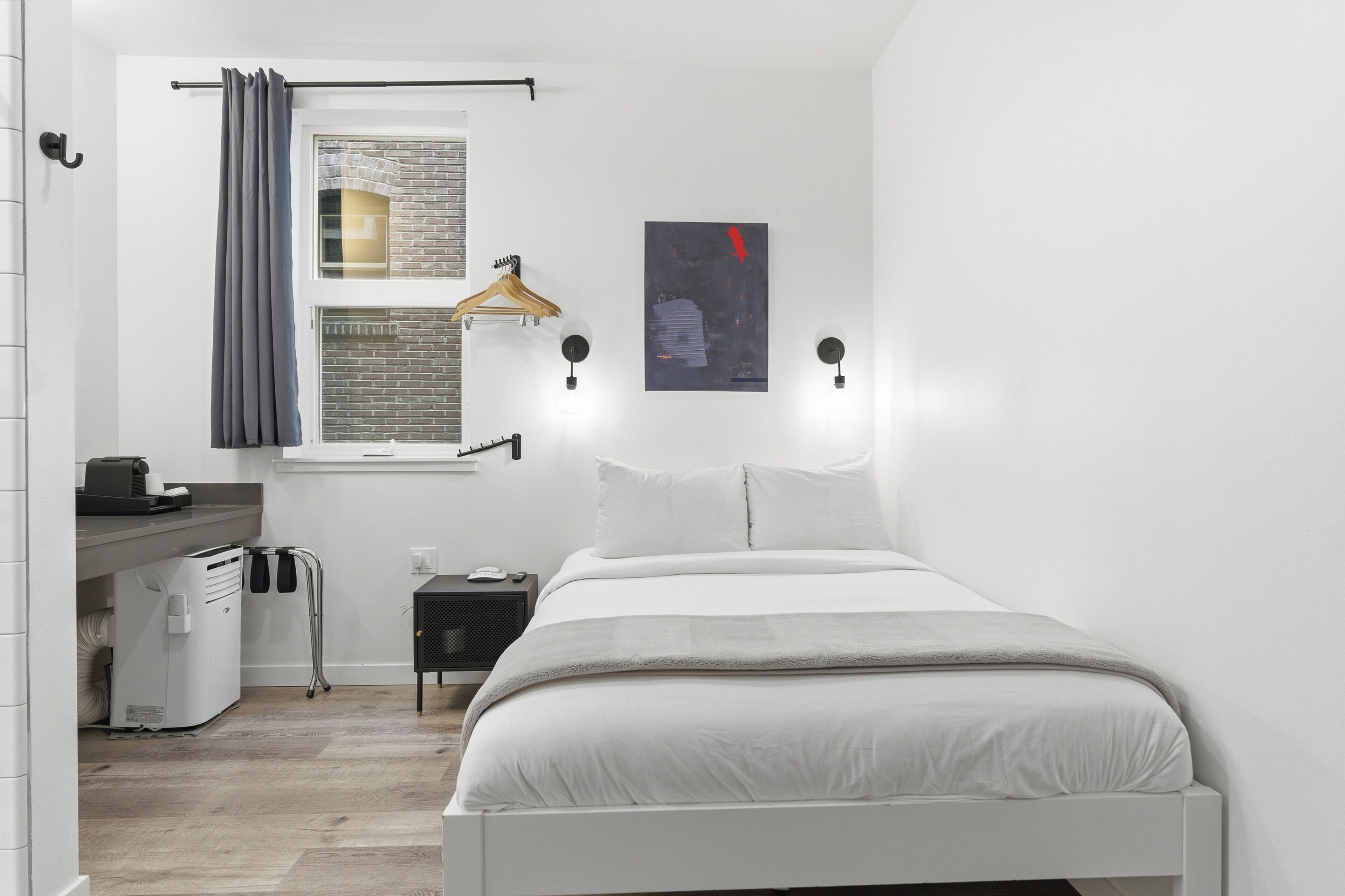 A bedroom with a bed, a window with a curtain, a wall-mounted light, a small bedside table, and a heater.