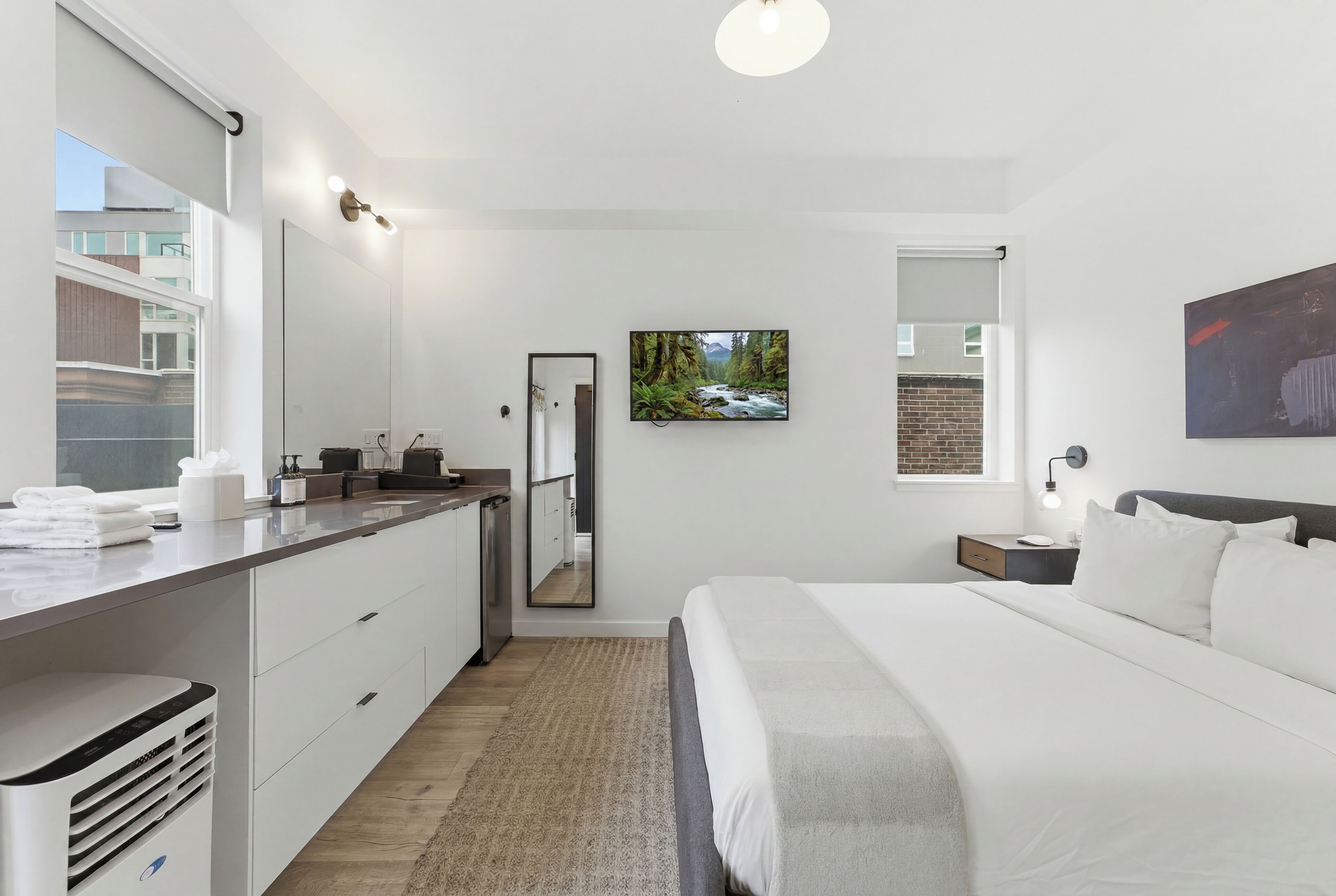 A modern hotel room with a bed, a desk, a mirror, a painting, and a view of the outside.