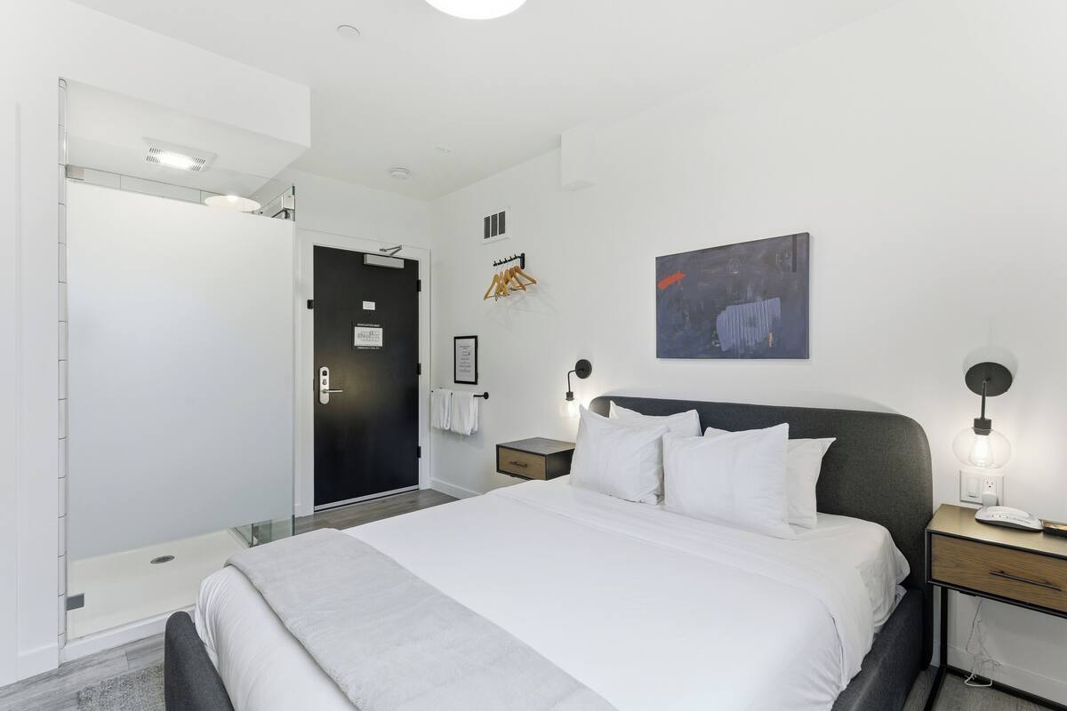 Une chambre d’hôtel moderne, équipée d’un lit, de tables de chevet, d’une œuvre d’art accrochée au mur et d’une porte donnant accès à une salle de bain.