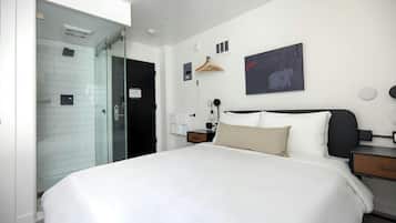 A hotel room with a large bed, a glass shower enclosure, a bedside table with a lamp, a wall-mounted artwork, and a wooden hanger.
