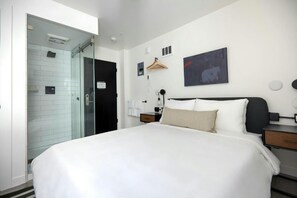 Habitación de hotel con una cama grande, cabina de ducha de vidrio, mesita de noche con lámpara y una obra de arte colgada en la pared.