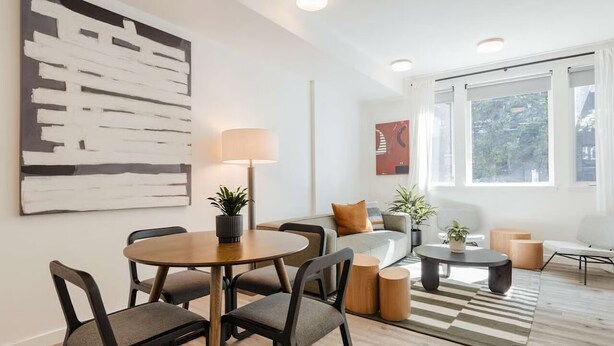 Un salon moderne avec une table de salle à manger ronde, des chaises, un canapé et une table basse. Un grand tableau abstrait orne le mur, ainsi qu’une plante en pot et une fenêtre avec des rideaux.