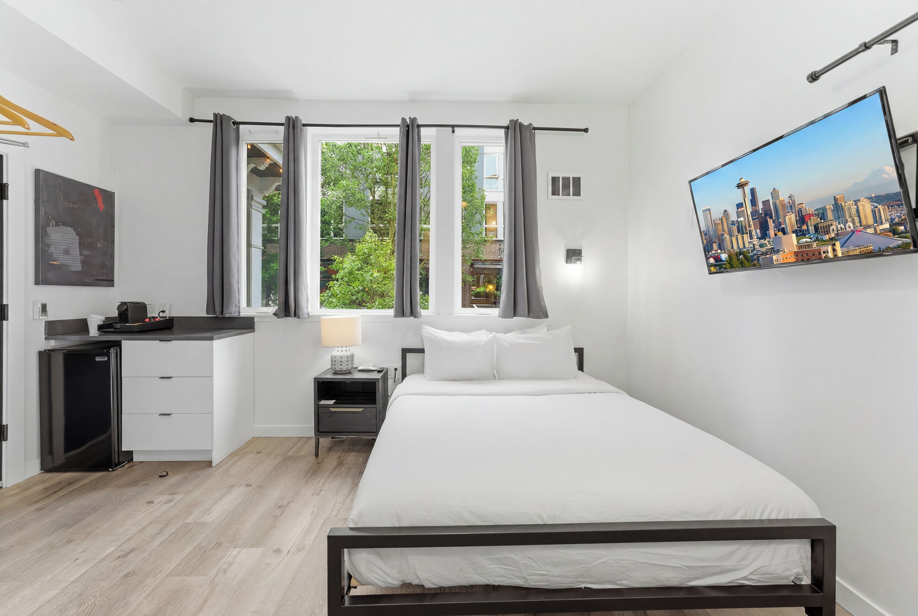 A modern hotel room with a bed, a desk, a small fridge, and a cityscape view.
