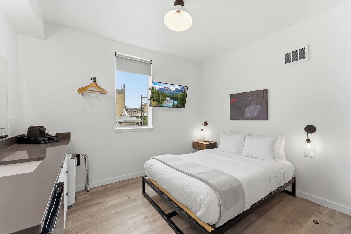 Une chambre moderne avec un lit, des tables de chevet, un luminaire suspendu, un téléviseur fixé au mur et une fenêtre donnant sur un beau paysage.