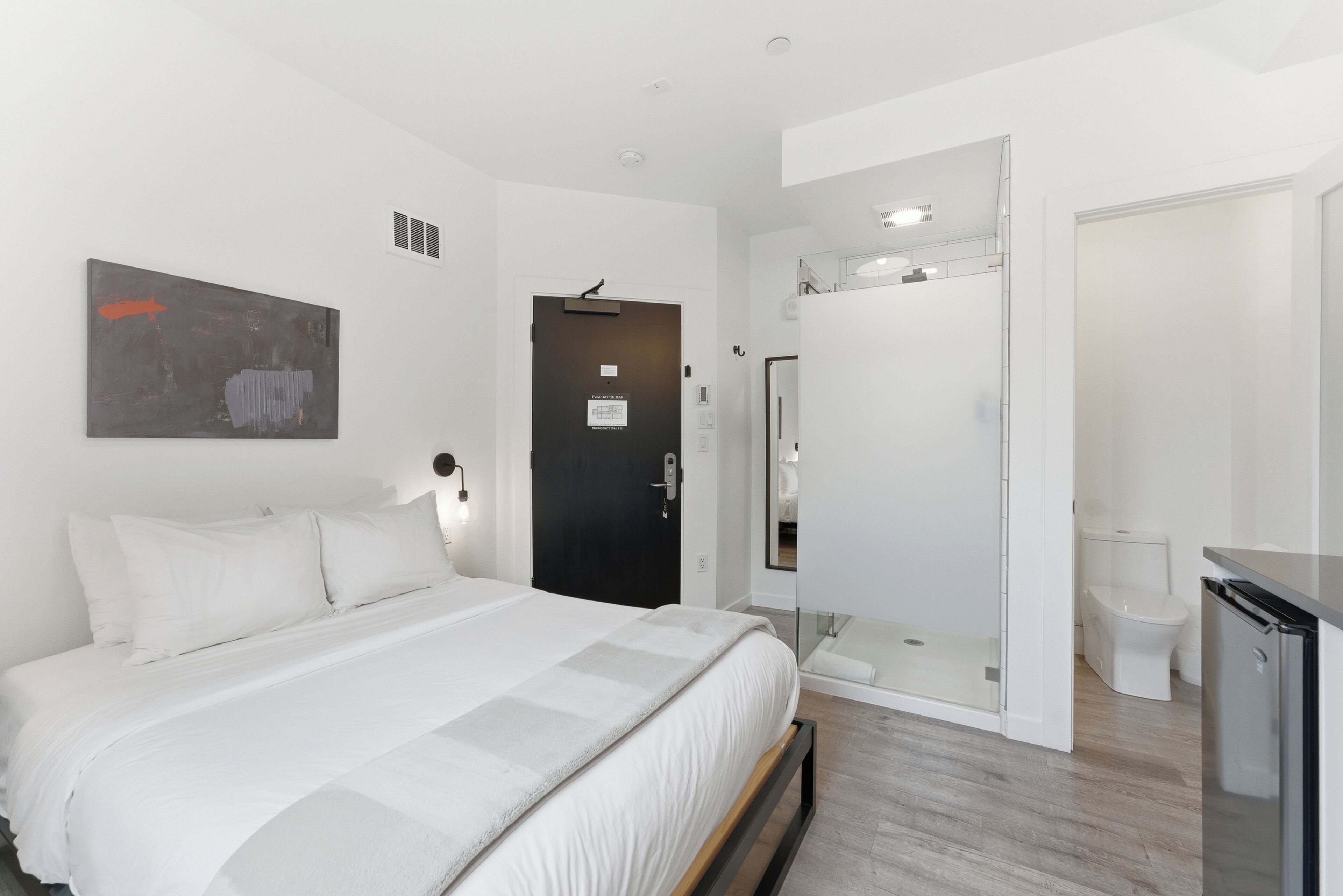 A modern hotel room with a bed, a shower, a toilet, and a small kitchenette.