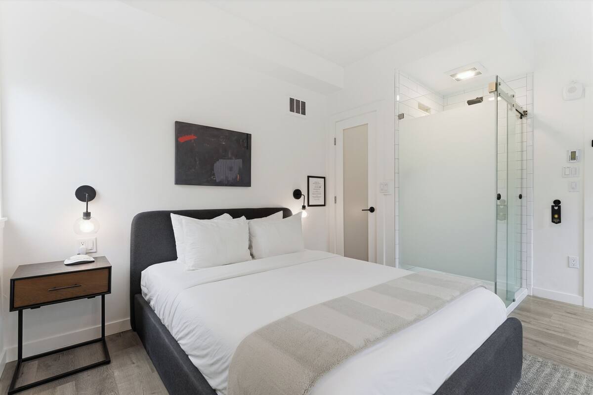 Une chambre moderne avec un lit, une table de chevet, une lampe, une œuvre d’art et une cabine de douche en verre.