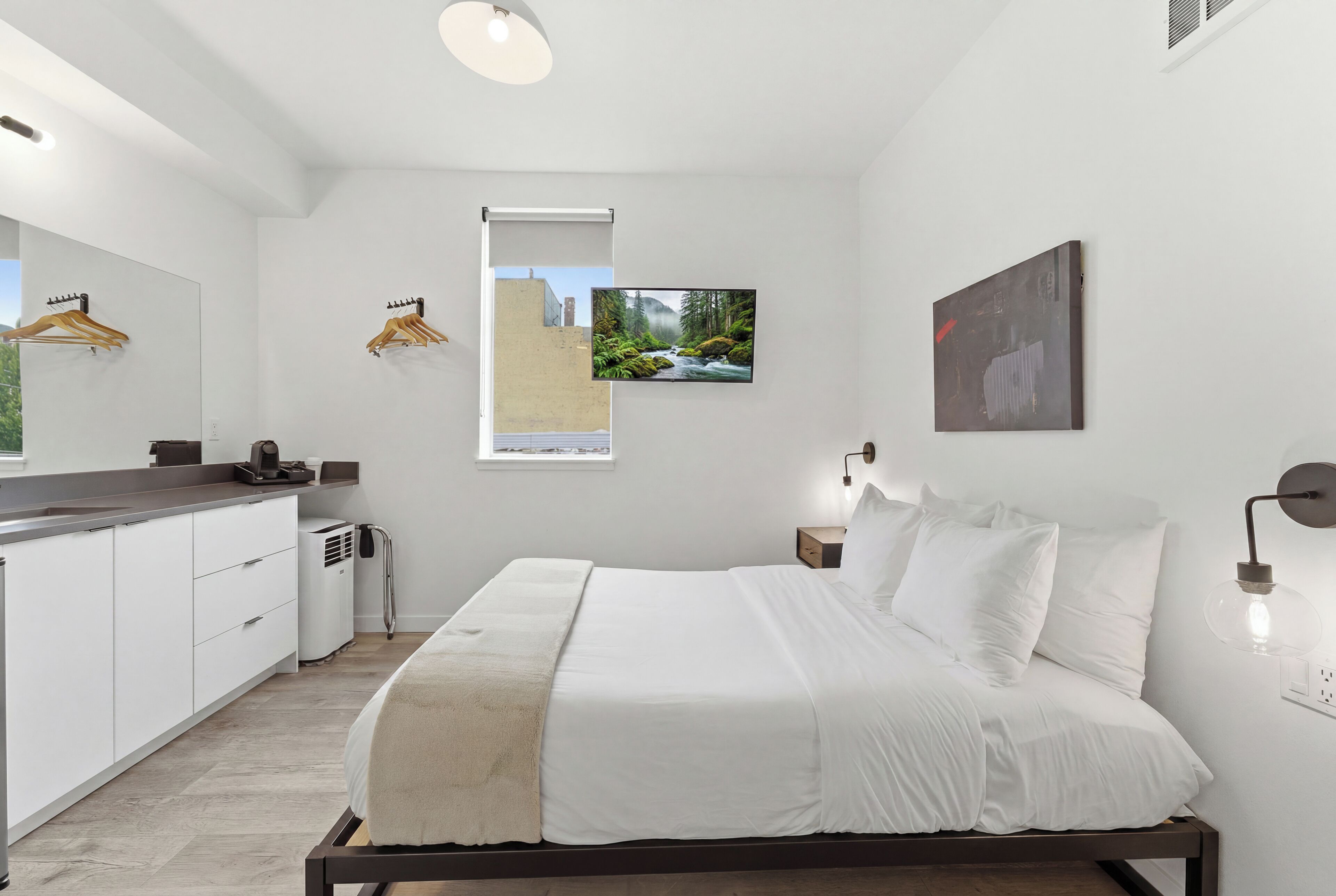 A bedroom with a bed, bedside tables, a window with a view, a wall-mounted artwork, and a kitchen area with cabinets and a countertop.