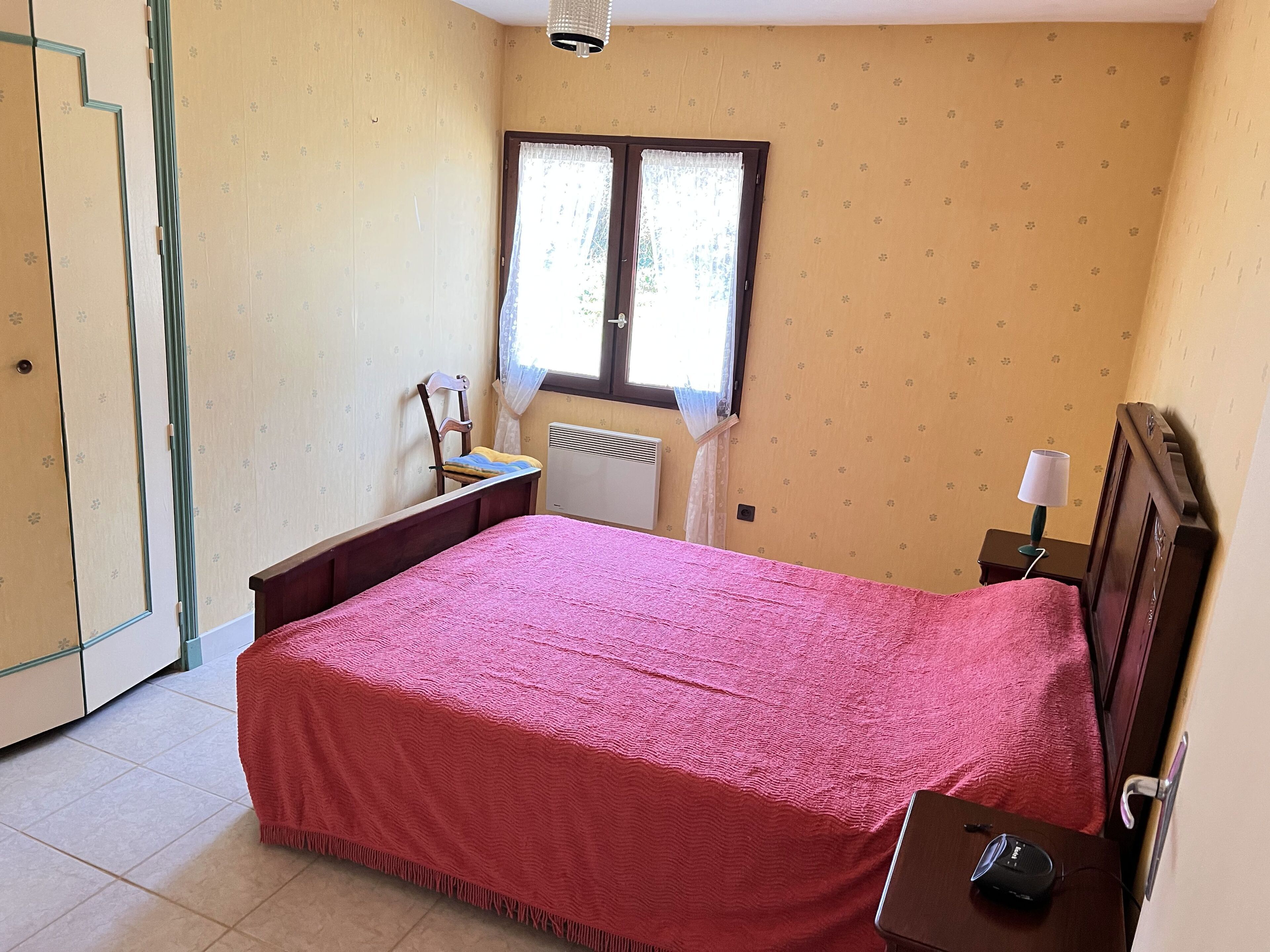 A bedroom with a bed, a nightstand, a chair, a window with curtains, and a radiator.