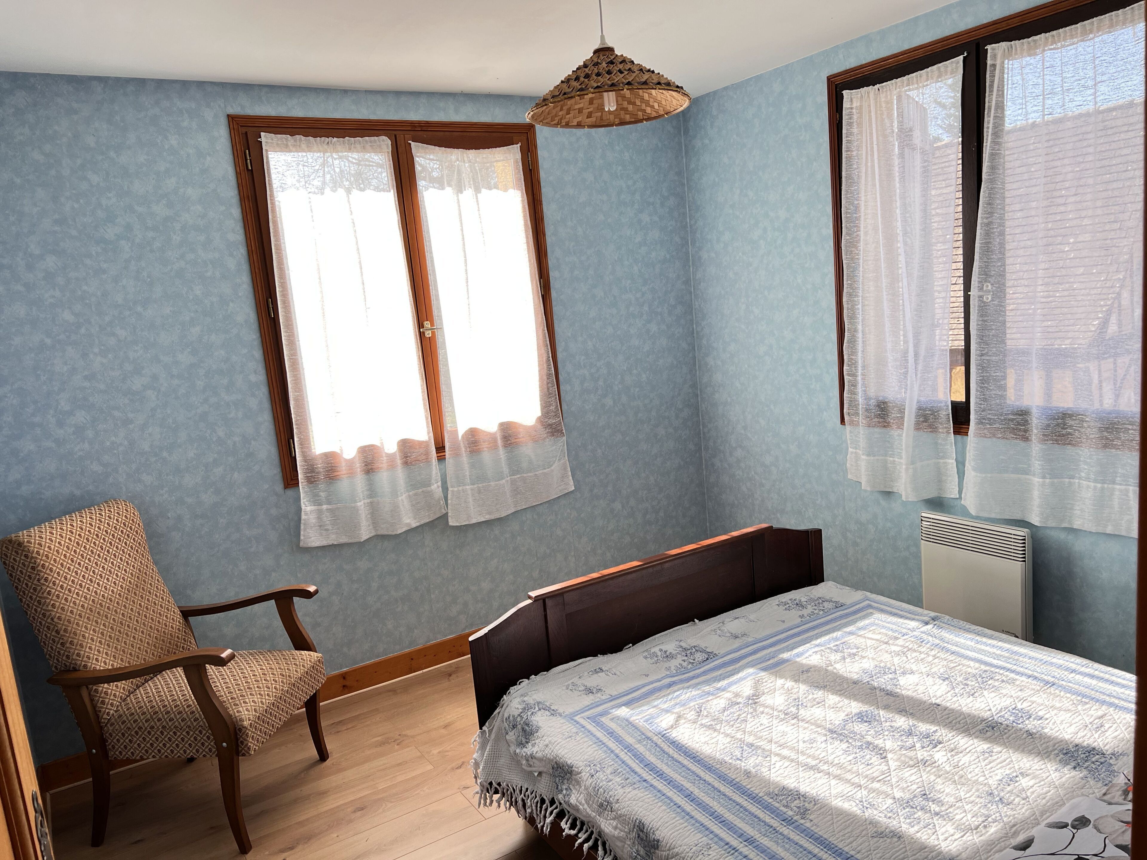 Une chambre avec un lit en bois, une chaise, une fenêtre avec des rideaux légers et un luminaire suspendu.