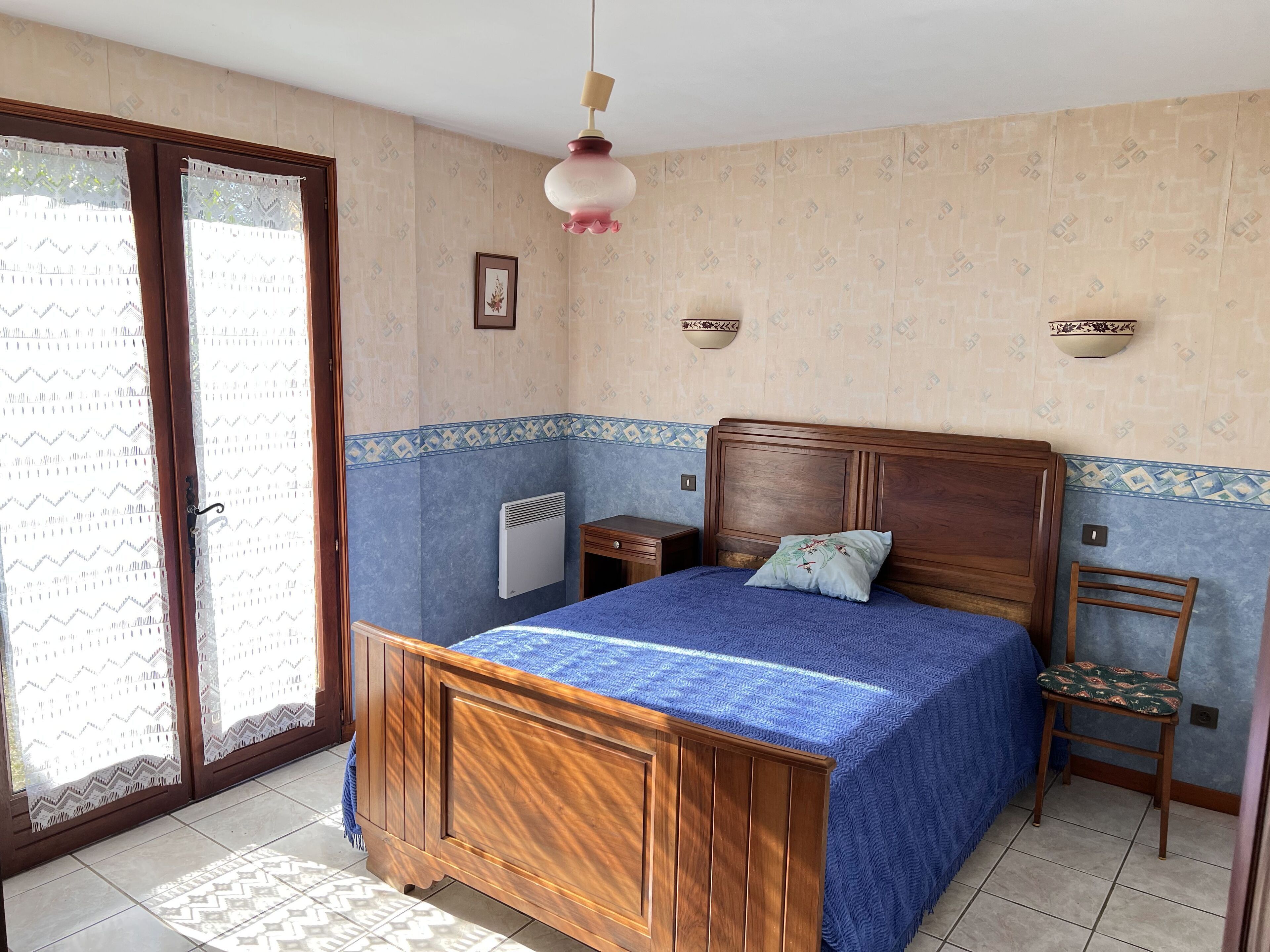 Una camera da letto con un letto di legno, una sedia, un comodino, un termosifone a parete e una finestra con tende.