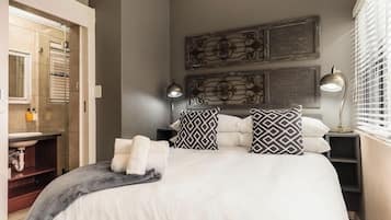 A modern bedroom with a large bed, two bedside tables, a wall-mounted light fixture, and a bathroom visible through an open door.