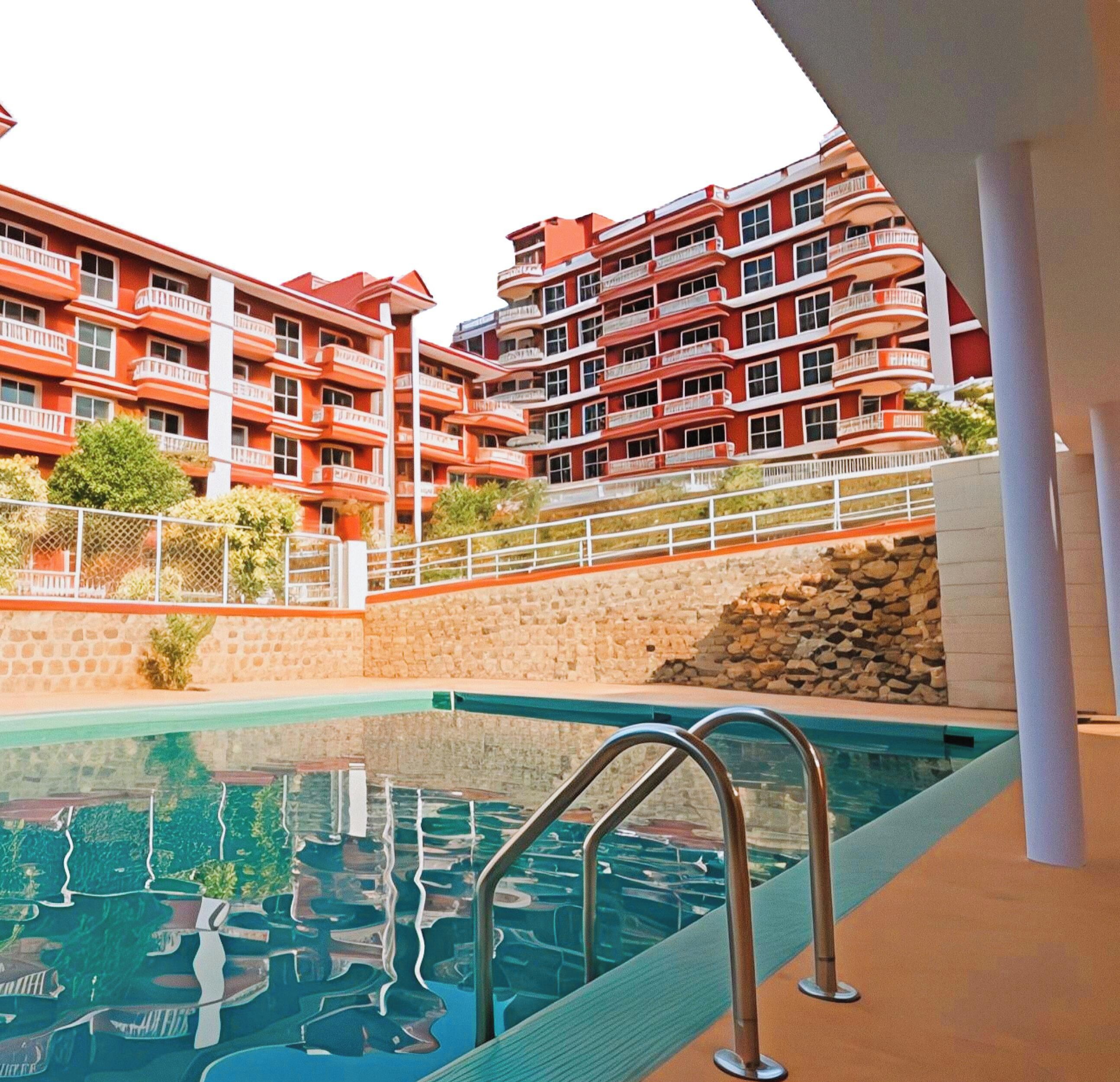 Une piscine entourée de rampes métalliques, elle-même ceinte d’un mur de pierre et bordée d’un immeuble doté de nombreux balcons.