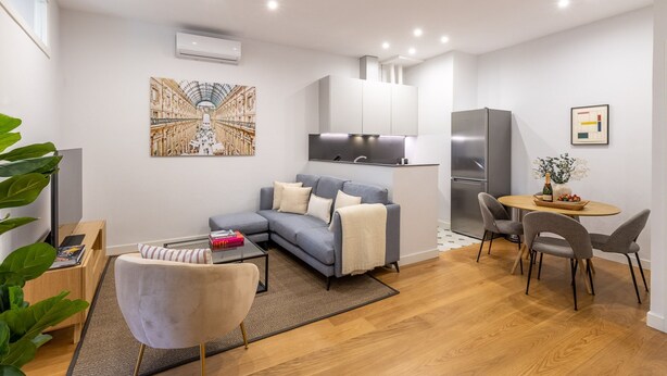 A modern living room with a grey sofa, a small round table with chairs, a kitchen area with a fridge, and an air conditioning unit. - Bright 2-Bed Apartment in Las Letras, Madrid (Madrid)