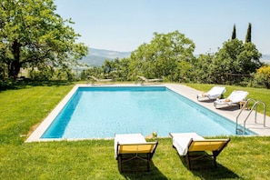 Una piscina rectangular con superficie de agua azul cristalina, rodeada por un césped bien cuidado y árboles.