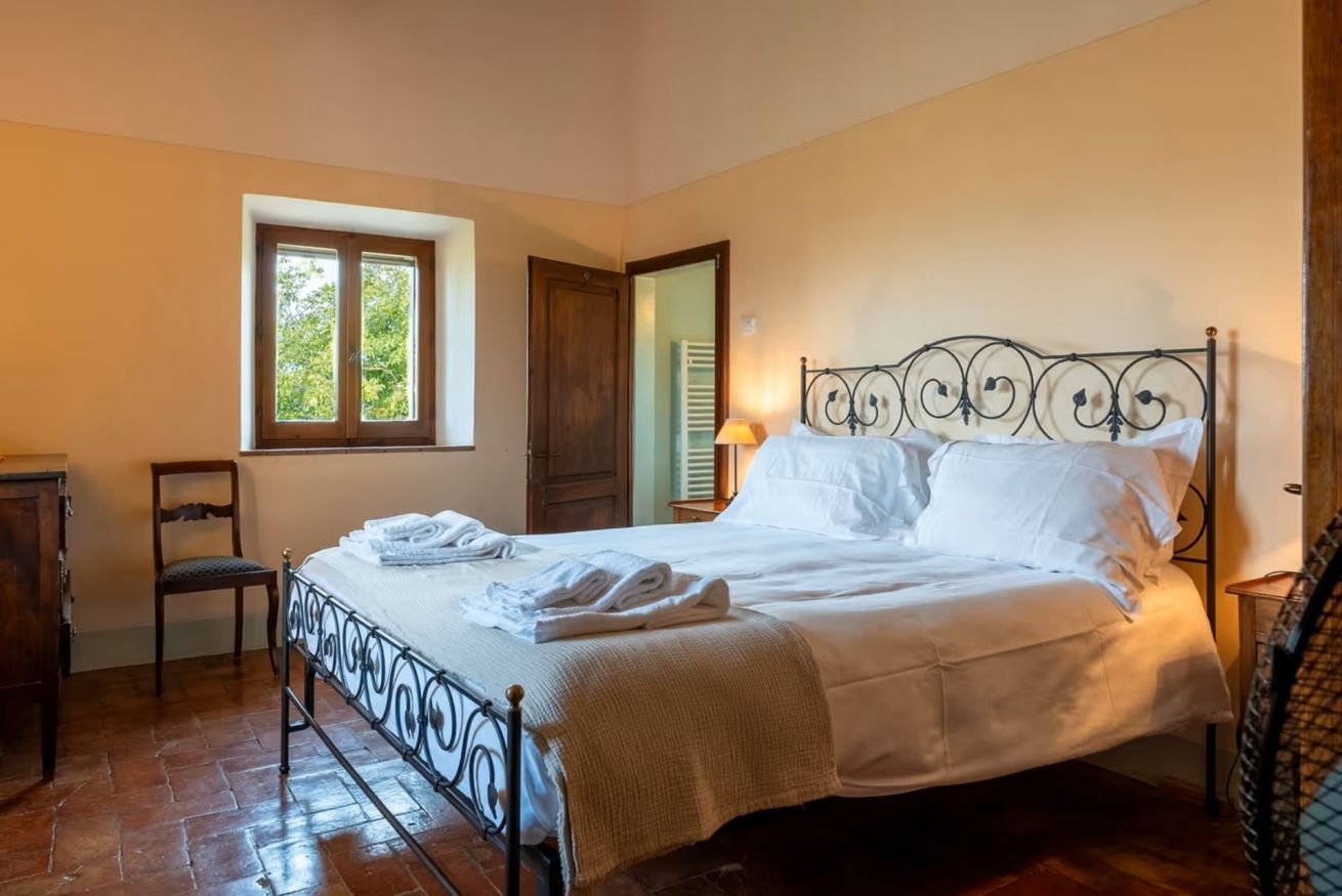 Un dormitorio con estructura de cama metálica, una silla de madera, un ventanal con vista a árboles y una puerta.