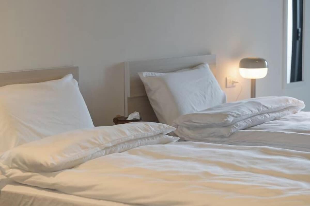 A neatly made bed with white bedding and pillows, a bedside lamp, and a window with a view.