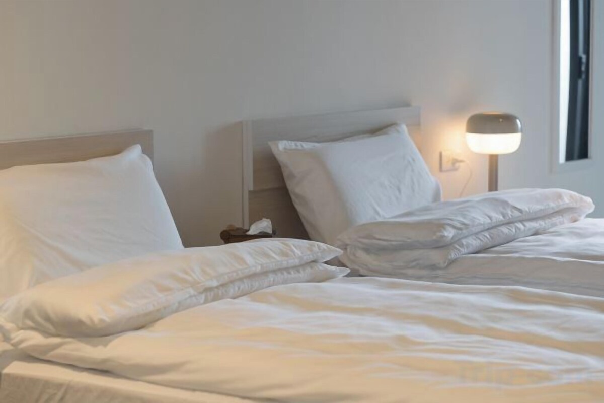 Cama bem arrumada com roupa branca e almofadas, luminária de cabeceira e mesa de apoio com um telefone.