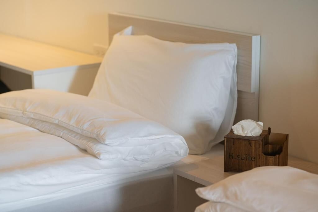 A neatly made bed with white linens and pillows, a bedside table with a tissue box labeled "H. suite".