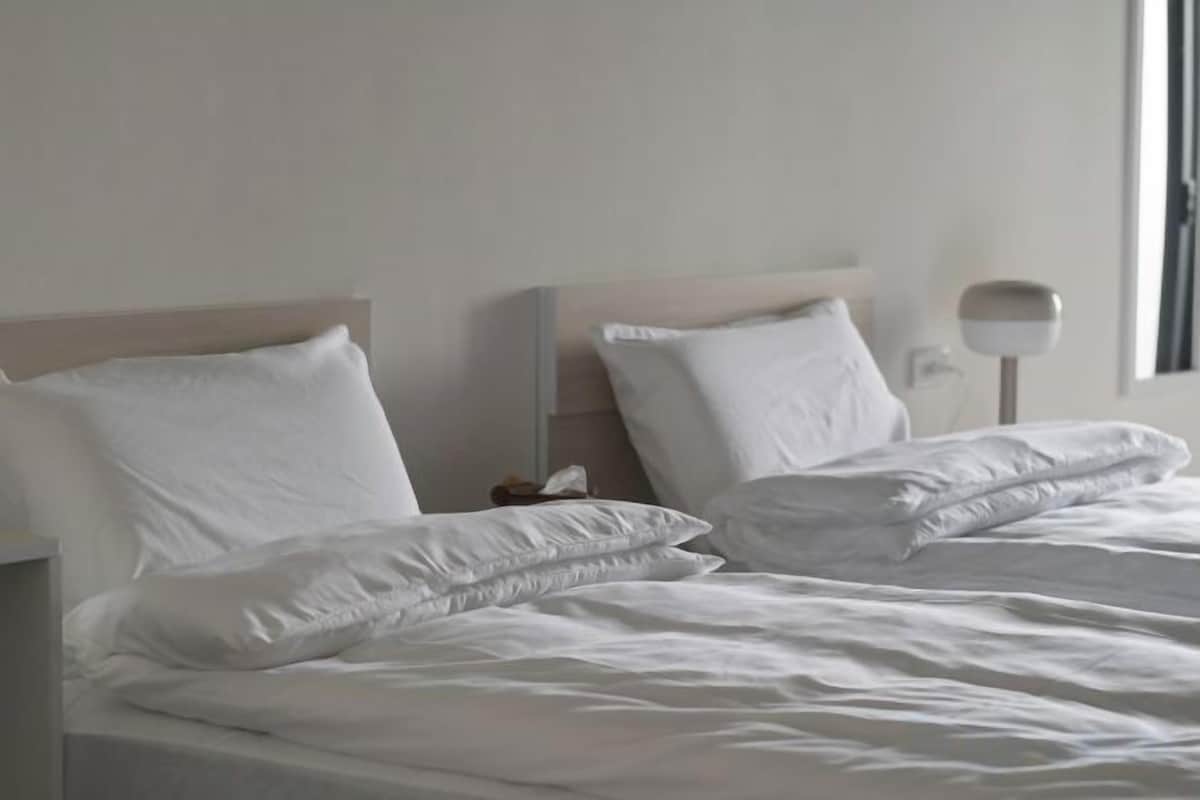Cama com roupa de cama e almofadas brancas, mesa de cabeceira e luminária na parede.