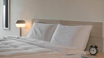 A neatly made bed with white linens, a bedside table with a clock, and a wall-mounted lamp.