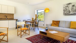 Ein modernes Wohnzimmer mit einer Couch, Couchtisch und Sesseln. Ein Esstisch mit Stühlen und ein Fenster mit Blick auf Grünflächen.