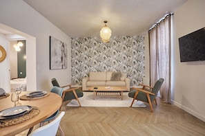 Un coin repas avec une table en bois dressée pour quatre personnes, un canapé et deux fauteuils. On peut voir un papier peint à motifs et un luminaire suspendu.