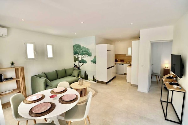 A modern apartment with a dining area, a living room with a green sofa, a kitchen, and a TV on a stand.