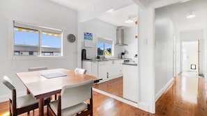 Cozinha moderna com armários brancos, área de jantar com mesa e cadeiras de madeira, e vista para o exterior pelas janelas.