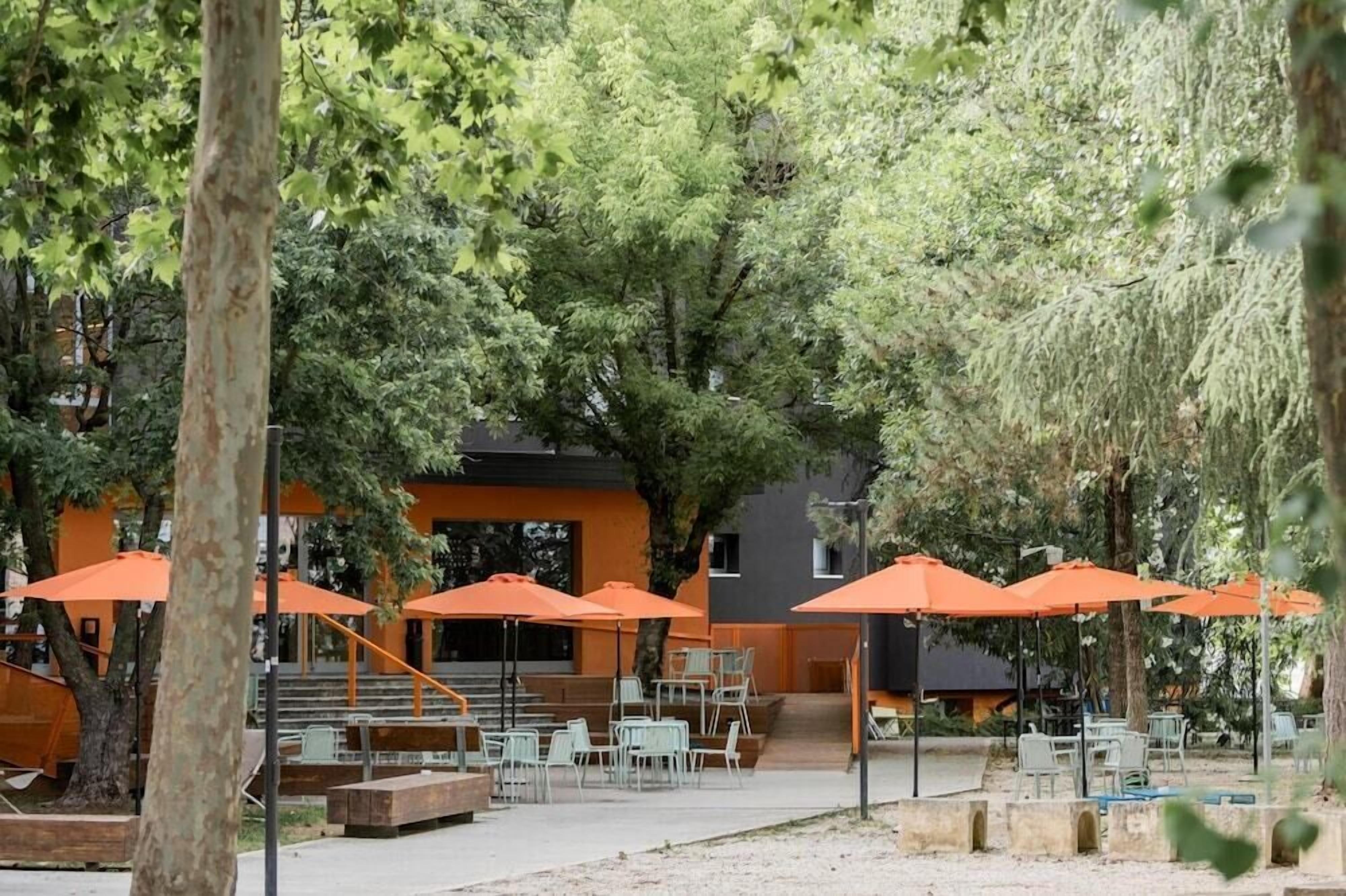 An outdoor seating area with orange umbrellas, white chairs, and a staircase leading to a building with a dark facade.