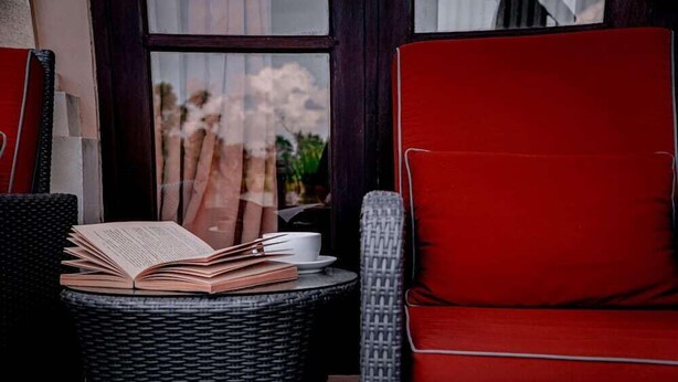 Una silla de mimbre con un cojín rojo, una mesita de mimbre con un libro abierto y una taza, y una ventana con vistas a árboles y nubes.