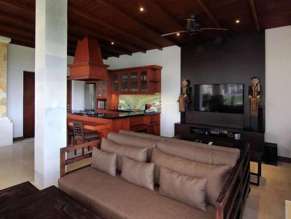 A modern living room with a sofa, a TV, and a kitchen area in the background. - Uma Stana Villa (Ubud)
