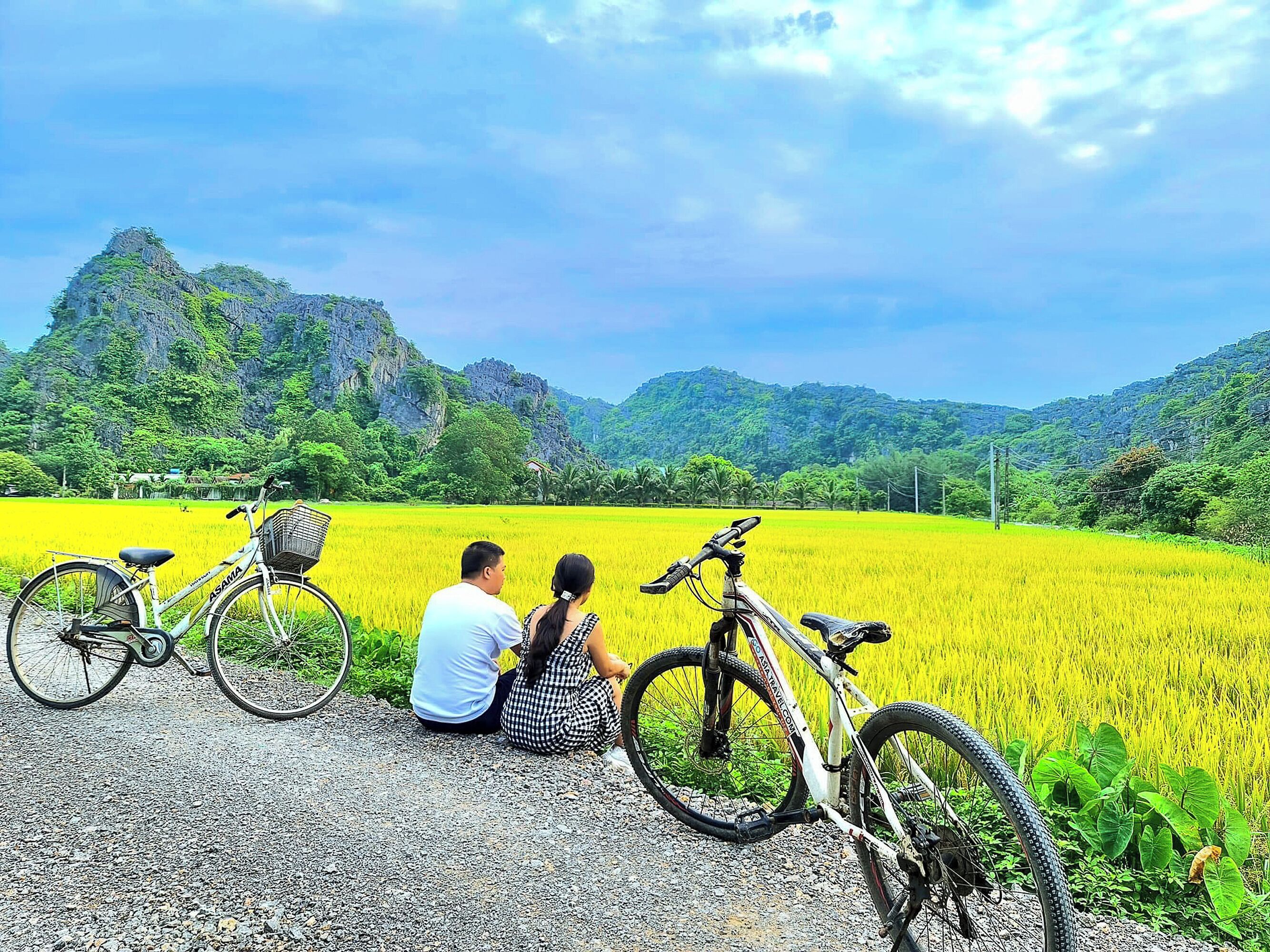 Una pareja sentada junto a una bicicleta en un campo, con montañas al fondo.