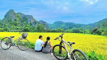 Una pareja sentada junto a una bicicleta en un campo, con montañas al fondo.