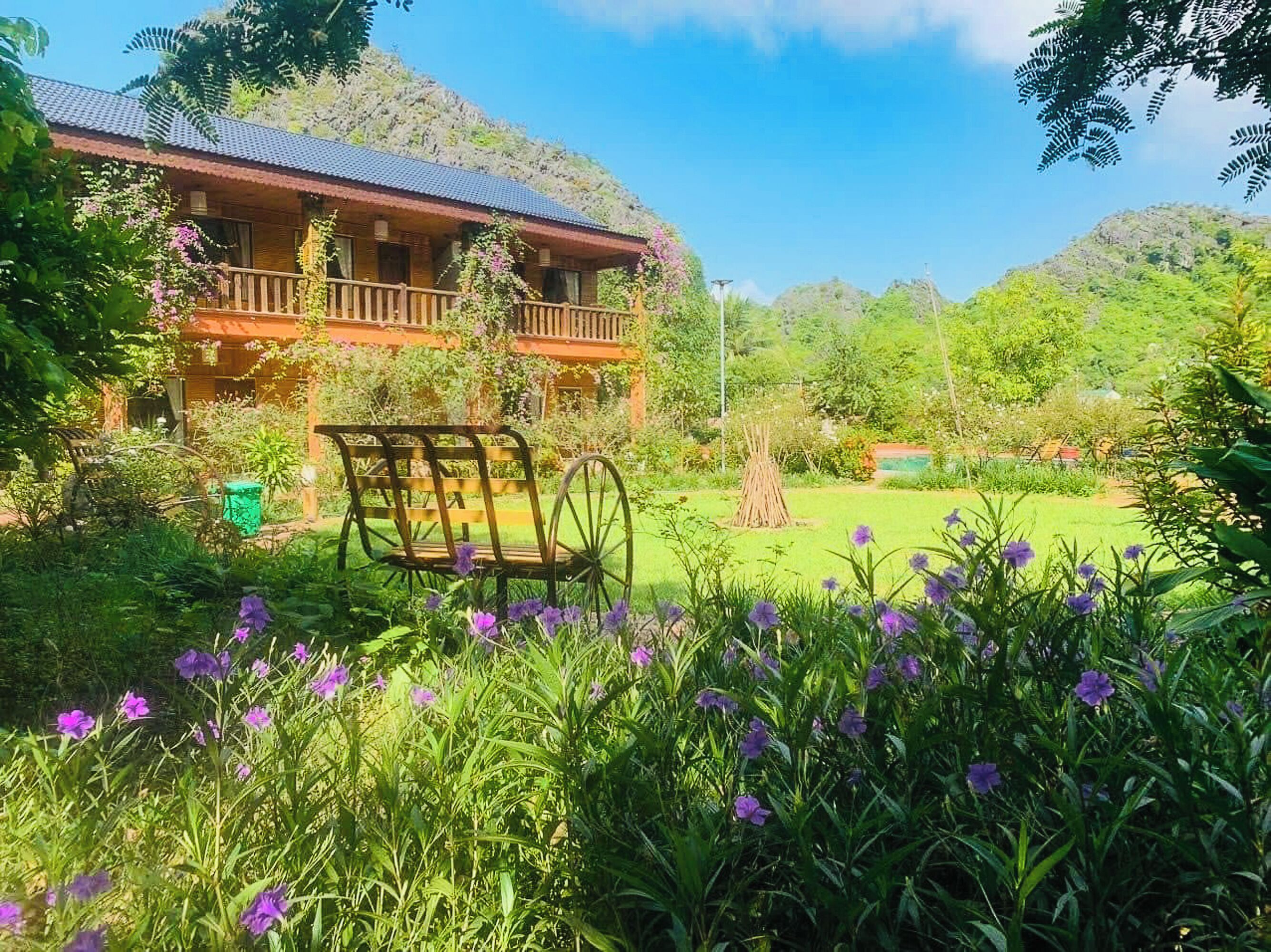 Una casa de madera rústica con balcón, rodeada de abundante follaje y flores moradas.