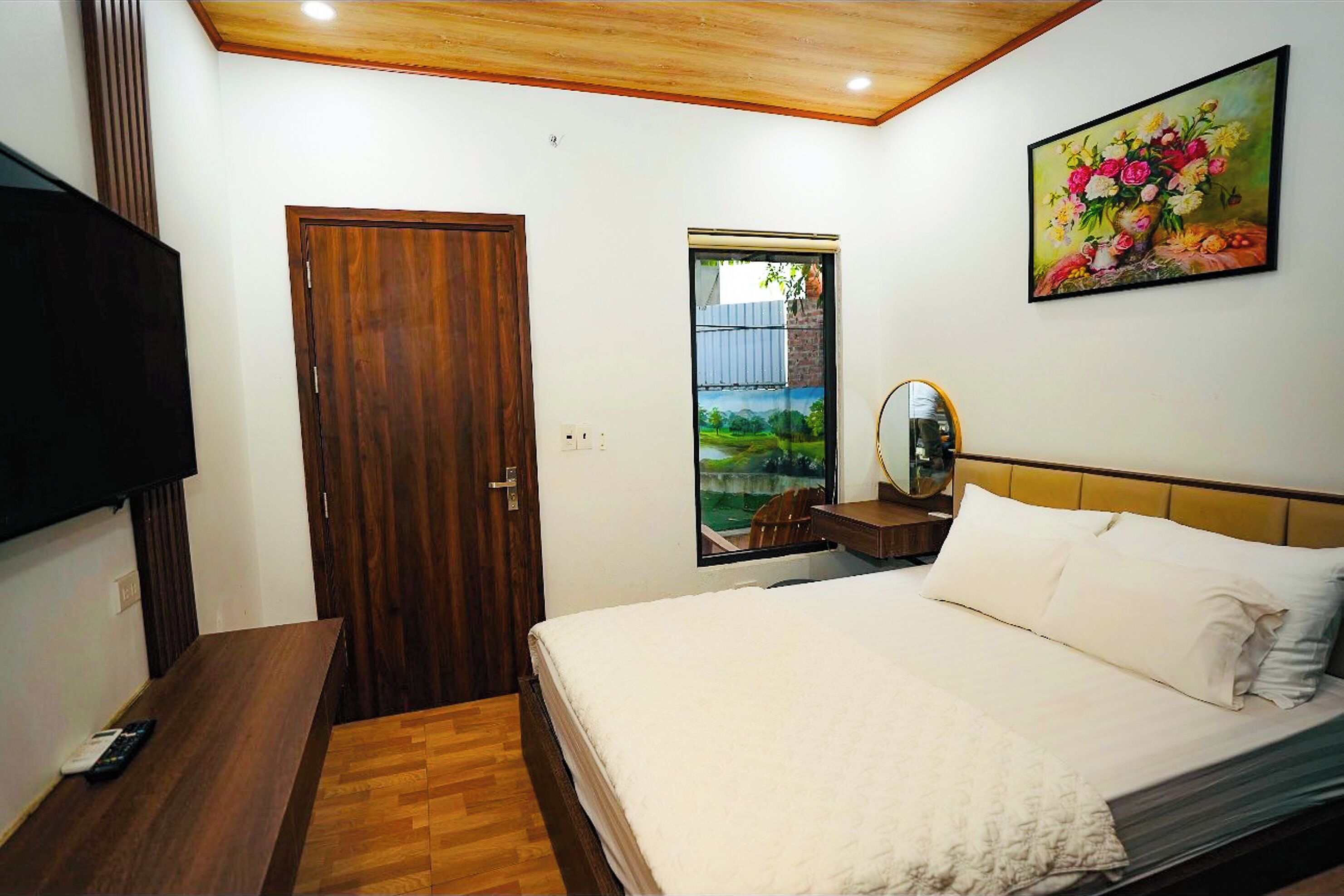 Un dormitorio con puerta de madera, ventana con vistas, cama con ropa blanca y un cuadro en la pared.