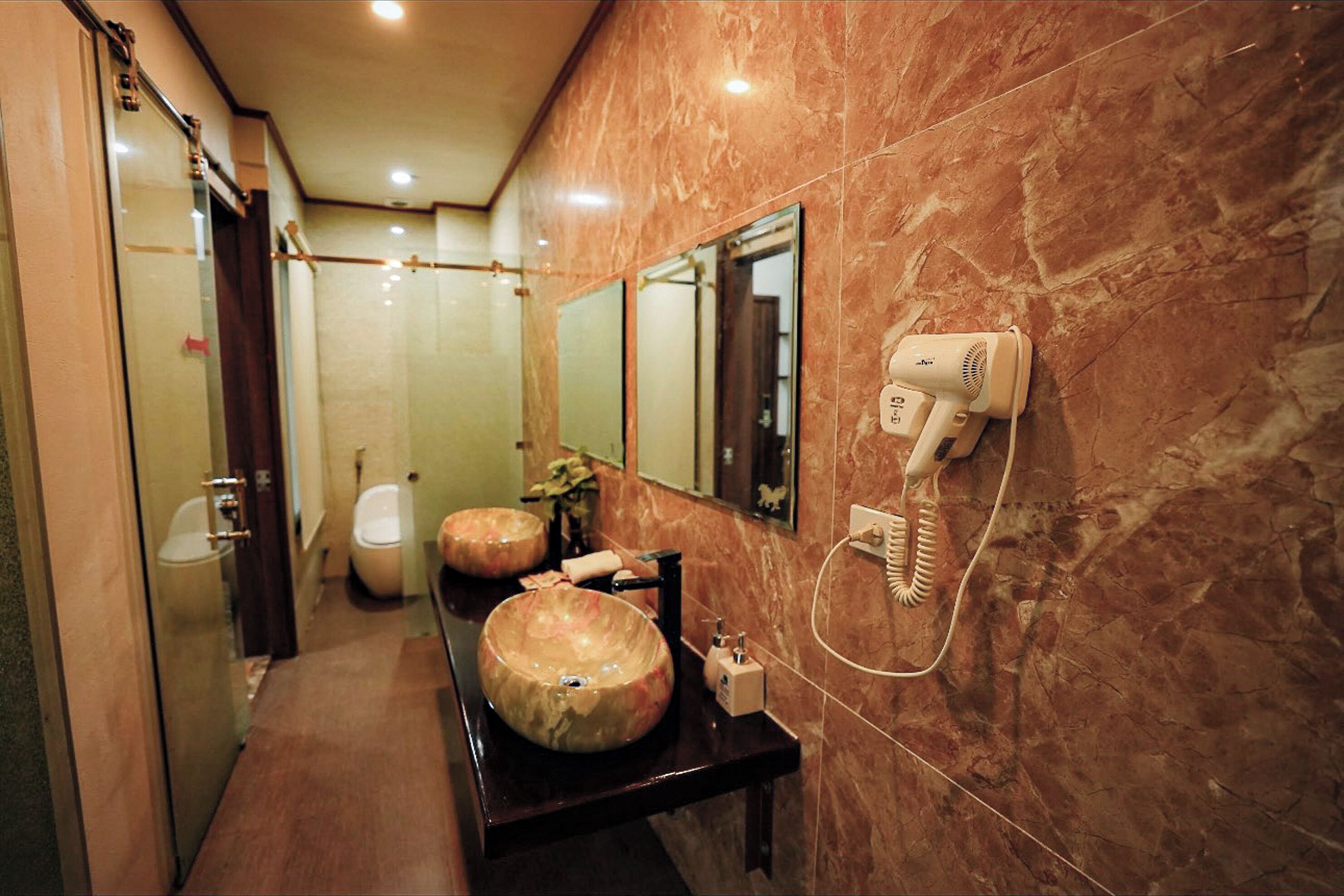 A bathroom with a marble wall, a large mirror, a sink with a vessel basin, a hairdryer on a wall-mounted charging station, and a toilet.