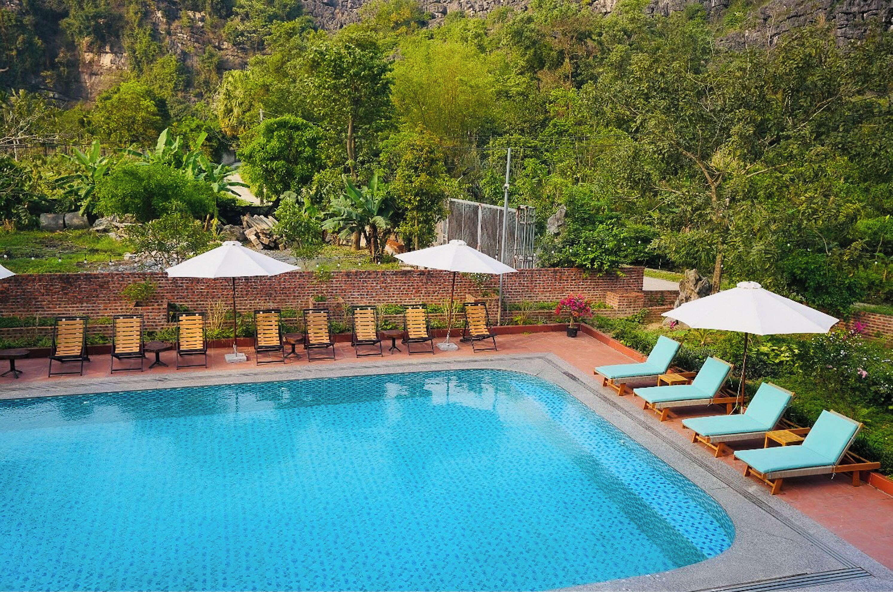 A swimming pool with lounge chairs and umbrellas, surrounded by greenery and a brick wall.