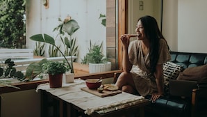 A person sitting on a couch in a room with a window, a table with a bowl and bread, and various potted plants. - Sasa Nor Lamour B B (Hengchun)