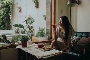 A person sitting on a couch in a room with a window, a table with a bowl and bread, and various potted plants. - Sasa Nor Lamour B B (Hengchun)