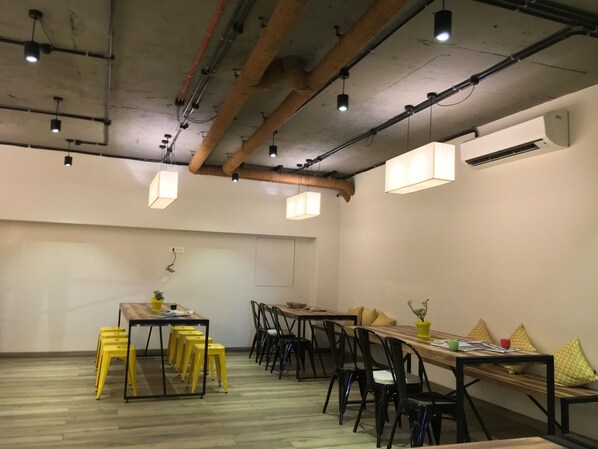Il s’agit d’une pièce spacieuse avec un plafond haut, des poutres apparentes et un éclairage de style industriel. On y trouve une longue table en bois, entourée de chaises, ainsi qu’une table plus petite avec des tabourets jaunes.