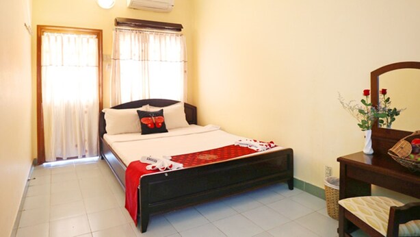 A bedroom with a bed, a bedside table, a mirror, and a window with curtains. - Hon Rom 1 Resort (Phan Thiet)
