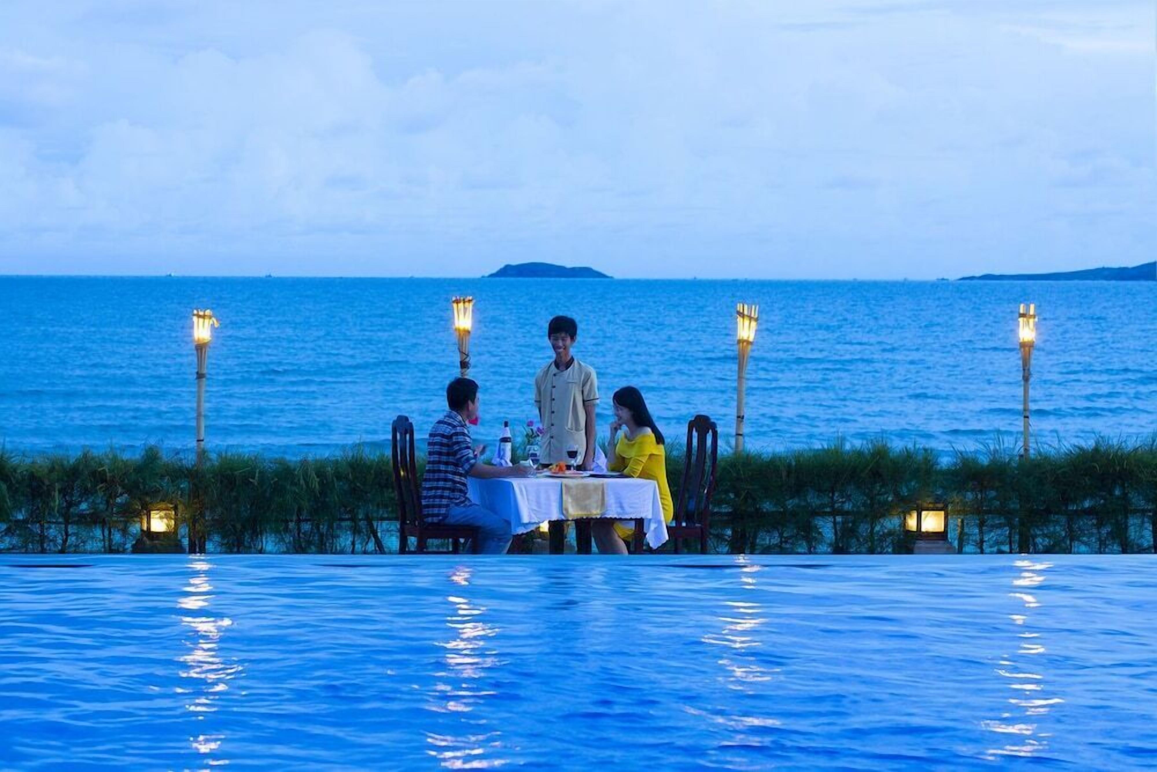 A dining table by a pool with a view of the sea, surrounded by chairs and illuminated by lanterns.