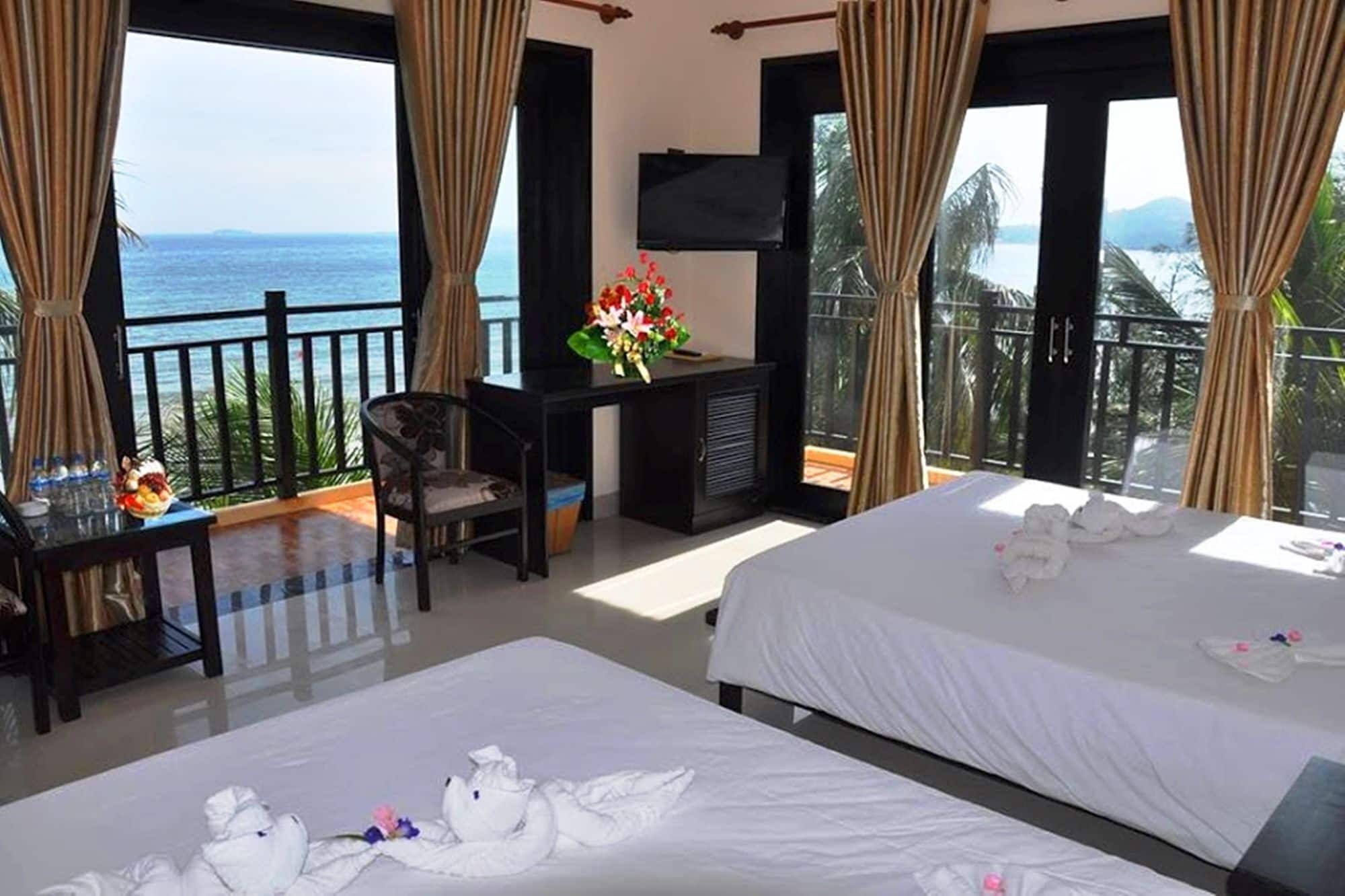 A hotel room with two beds, a desk, a TV, and a balcony with a view of the sea.
